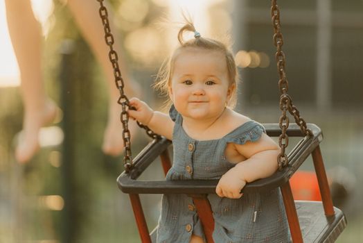 A happy toddler baby girl in a dress on a swing on the warm summer evening