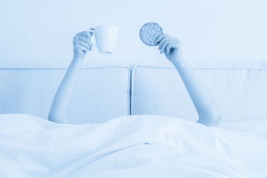 Female hands holding cup of coffee and cookie above blanket in bed. Woman having breakfast in the morning