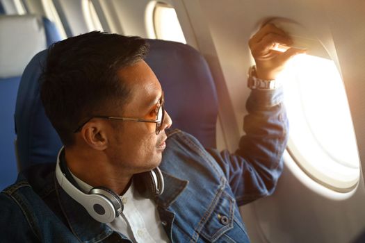 Hipster male traveller opening airplane window and watching outside landscape during beautiful sunrise