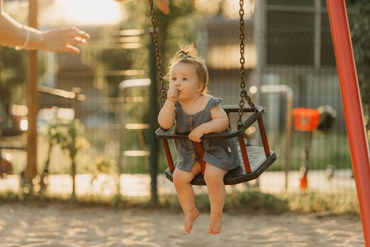 Toddler in a dress is sucking her thumb on a swing on the warm summer evening
