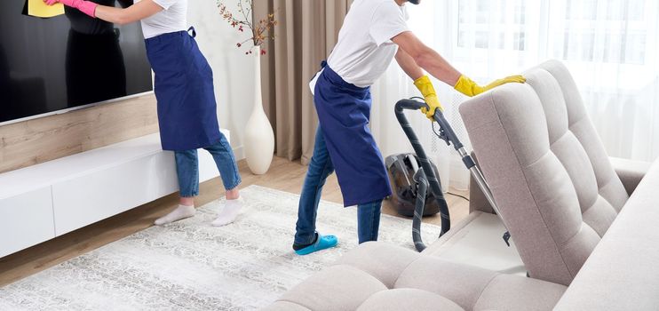 Professional cleaners in blue uniform washing floor and wiping dust from the furniture in the living room of the apartment. Cleaning service concept