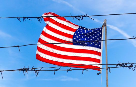 Barbed wire fence in front of Flag of United States of America waving in the wind with blue sky background