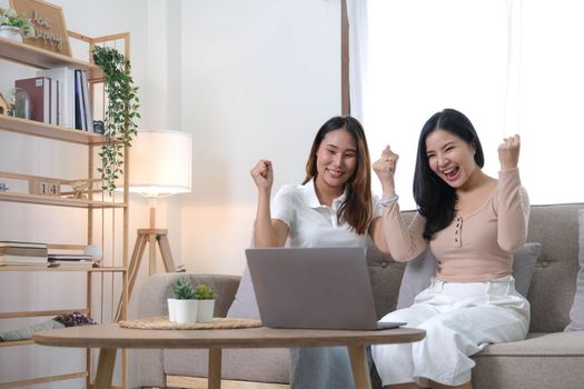 Two happy Asian women best friends in casual wear laughing while working with laptop at home in living room