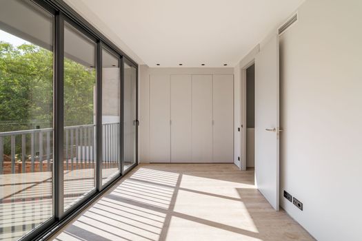 Sunlight illuminates the room, creating shadows on the floor. New room in a refurbished apartment with wooden floor, wardrobe and big windows.