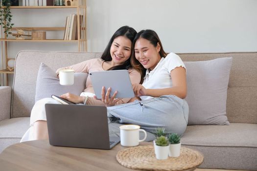 Two happy Asian women best friends in casual wear laughing while working with tablet at home in living room
