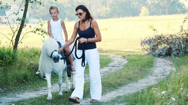 Mom and daughter are walking around the field, daughter is riding a pony, mother is holding a pony for a bridle. Cheerful, happy family vacation. Outdoors, in summer, near the forest. High quality photo