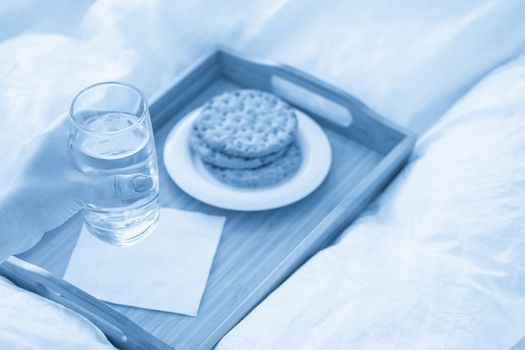Female hand holding glass of water over tray with crackers on bed