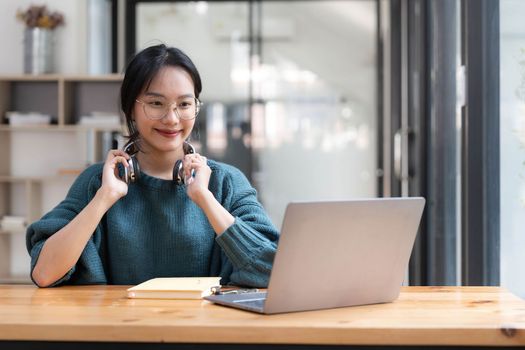 happy young asian ethnicity female manager wearing wireless headphones, looking at laptop screen, holding pleasant conversation with partners clients online, working remotely at workplace.