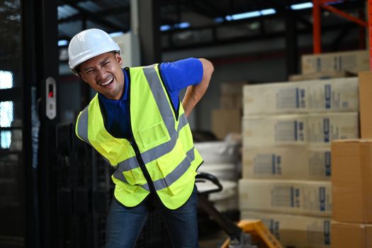 Strong asian man worker wearing hardhat and vests pulling pallet truck walking through rows of storage racks with merchandise