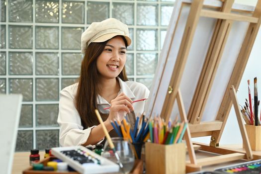 Smiling female painter sitting in front of canvas and painting picture with watercolor. Leisure activity concept