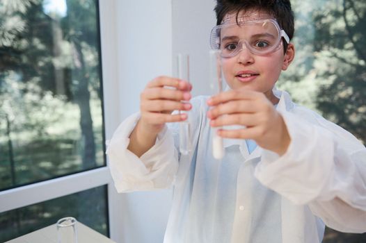 Smart schoolboy with safety goggles, in lab coat, looking at test tubes, fascinated by the going on chemical reaction