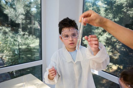 Teenage schoolboy in lab coat and goggles, attentively watches and analyzes a solution in test tube, at chemistry class
