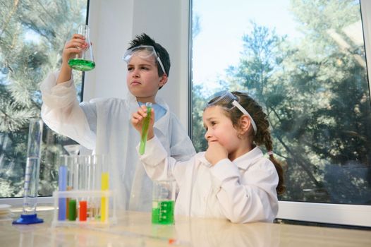 Two smart children, teenage boy and preschooler girl conducting chemical experiments in school science laboratory