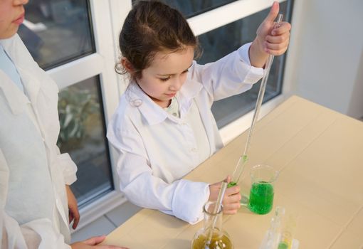 Smart little girl in lab coat, filling a graduated pipette with green chemical liquid in the school chemistry laboratory