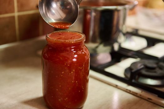 Selective focus on a sterilized canning can and housewife pouring freshly brewed tomato juice from a saucepan into it