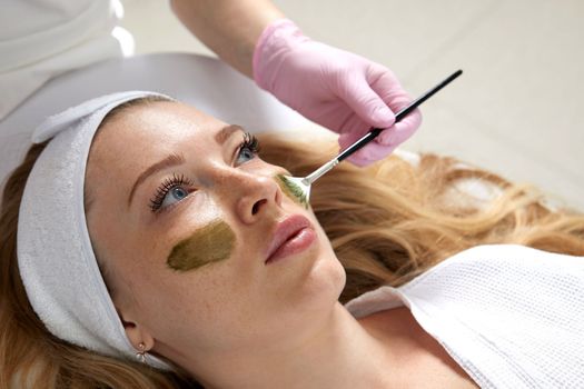 Cosmetologist applying a clay mask to the face of beautiful woman. Spa treatment and face care in the beauty salon
