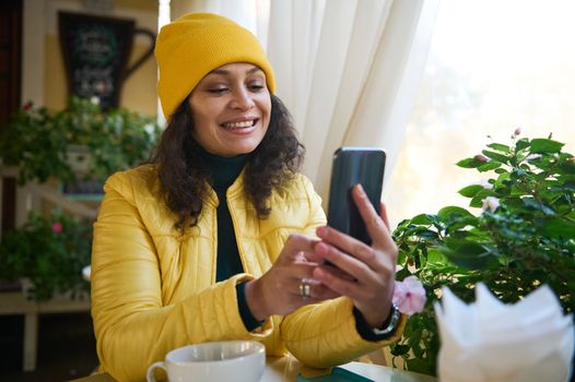 Charming multi-ethnic woman in bright yellow clothes, smiling a toothy smile while having a video call on her smartphone
