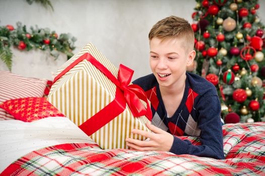 A cheerful teenager boy opens a Christmas gift. Cheerful Teenager lies in bed with a christmas present in his hands