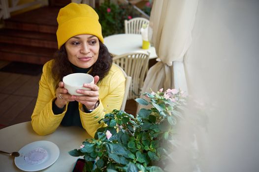 Stunning millennial brunette woman in a bright yellow hat and jacket, relaxing in a cafe, taking sip of hot aroma coffee