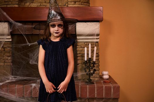 Little child girl in witch costume, stands against a cobweb covered fireplace and candles, ready for Halloween party