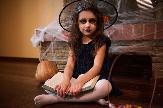 Little girl witch in wizard hat, with a book of sorcery, looks at camera sitting by a fireplace covered with cobwebs