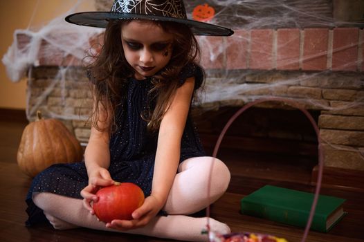 Little girl sorceress playing with a bright orange pumpkin, sitting on the floor near a spell book, against a fireplace