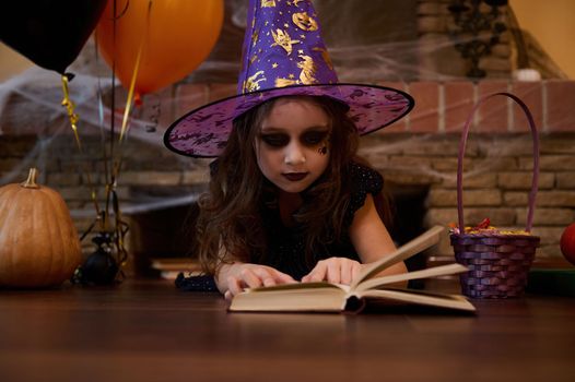 Little girl looking like a witch in wizard hat, lying on the floor, studying a spell book, surrounded by Halloween decor