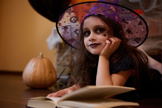 Little girl with a black spider painted on her cheek, looking like sorceress in wizard hat, smiles at camera. Halloween
