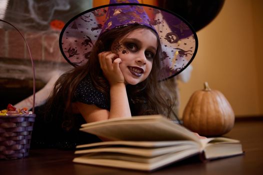 Inspired little girl witch in wizard hat, cutely smiles looking mysteriously at camera while reading sorcery spell book