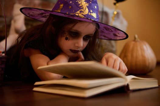 Close-up little witch, enchantress in wizard hat reading sorcery and spell magic book surrounded by Halloween atmosphere