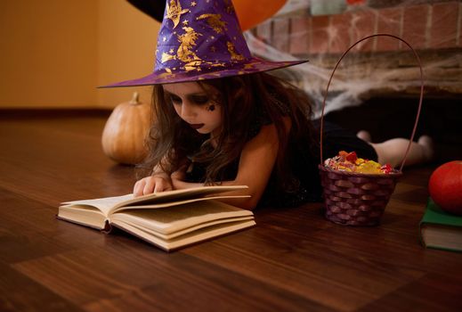 Little child girl focused on reading enchantery literature and sorcery spell book, surrounded by Halloween decor at home