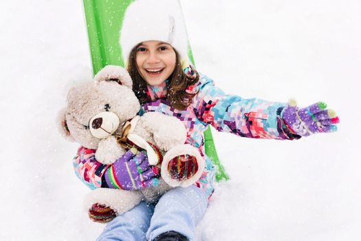 Portrait of child with toy teddy bear sits on a sled and looks at the winter snowy mountains.Winter vacantion. Christmas celebration and winter holidays. Winter fun and outdoor activities with kids