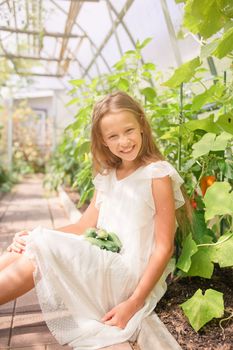 Adorable girl harvesting cucumbers and tomatoes in greenhouse. Portrait of kid with basket with vegetables