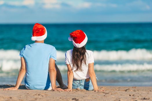 Young romantic couple in red Santa hats sitting on tropical white sand beach celebrating Christmas