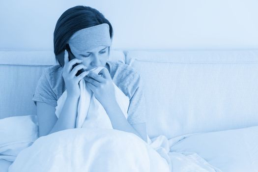 Sad woman wearing sleeping mask using smartphone as she sits in bed covered with duvet