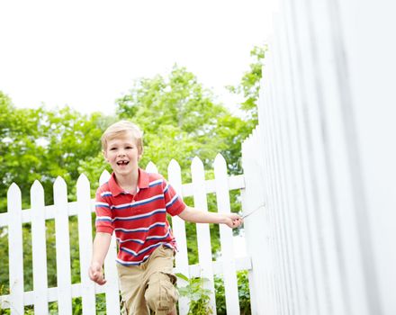 Oh to be young again. A cute young boy running a stick along a white picket fence in his backyard.
