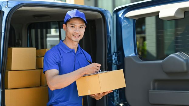 Smiling delivery man sitting in open delivery van and checking checking address details of client on clipboard