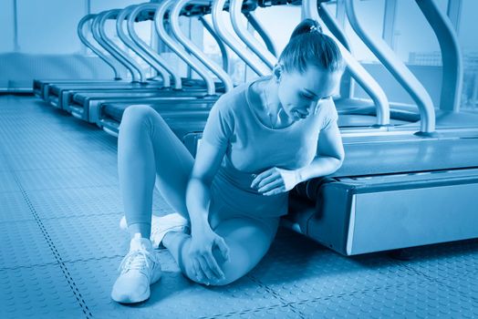 Young woman in sportswear having ache in knee while training in gym, Girl sitting on a floor touching her knee in pain