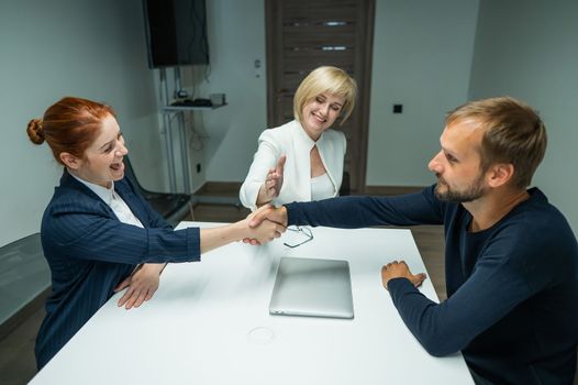 Blond, red-haired woman and bearded man dressed in suits in the office. Business people shake hands making a deal in a conference room.