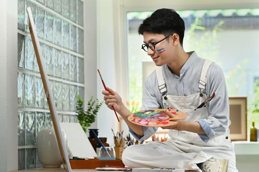 Smiling man artist sitting in front of canvas and painting picture with watercolor. Art and leisure activity concept