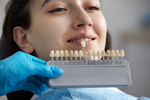 Dentist choosing color of tooth enamel for patient. Dentist applying sample from tooth enamel scale to caucasian female teeth