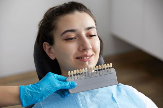 Dentist choosing color of tooth enamel for patient. Dentist applying sample from tooth enamel scale to caucasian female patient teeth