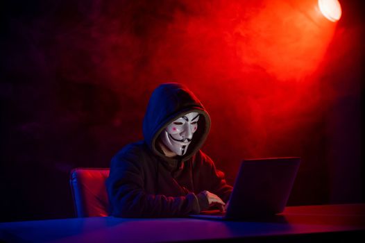 June 5, 2022 Novosibirsk, Russia: Anonymous in a hood is typing on a laptop in the dark in red-blue smoke.