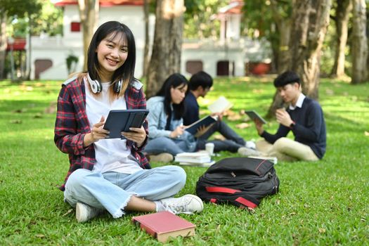 Cheerful asian female college or university student using digital tablet on green grass in the campus