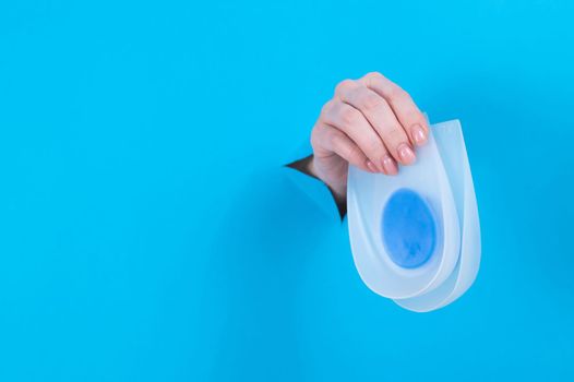 Woman holding orthopedic silicone heel pads. A woman's hand sticks out of a hole in a cardboard blue background.