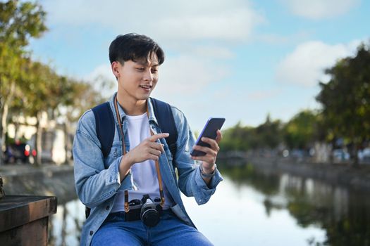 Asian male traveller searching direction on smart phone while sitting on bridge bench near river