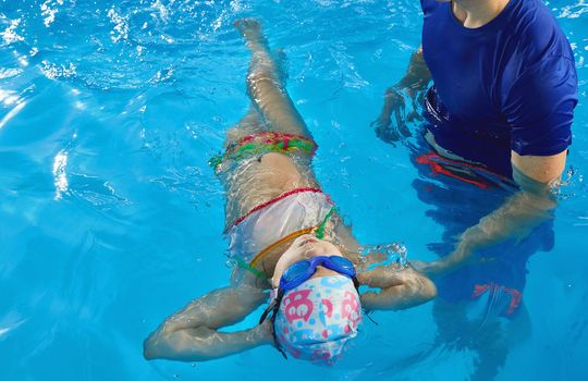 Trainer teaching preschool girl how to swim on her back in indoor pool