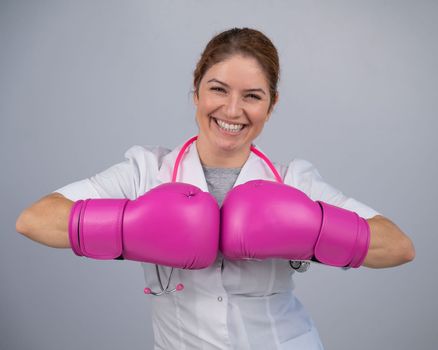 Smiling female doctor in pink boxing gloves with a pink ribbon on a gray background. Fight against breast cancer.