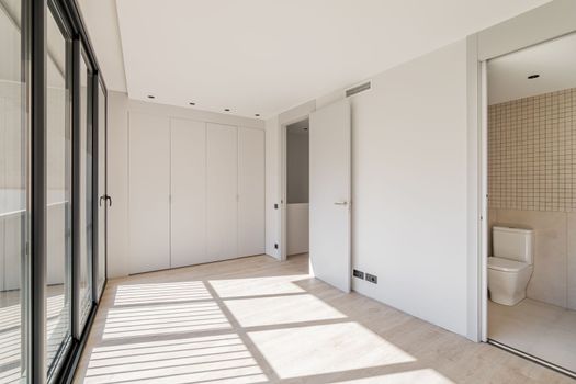 An empty room with built-in wardrobe in entire wall. With separate rooms for toilet and laundry. Room with large windows throughout wall through which bright sun illuminates spacious room.
