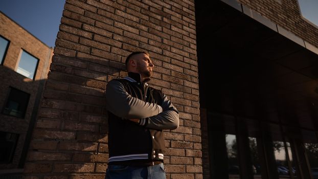 Caucasian bearded man in a bomber jacket leaned against a brick wall.